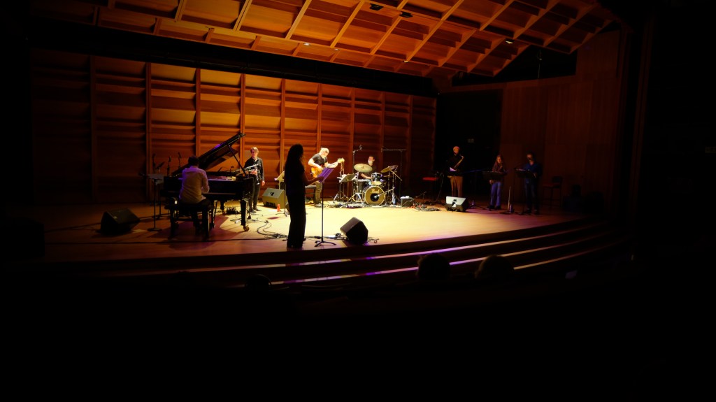A live music performance in a wooden auditorium with a piano, drums, and several musicians on stage.